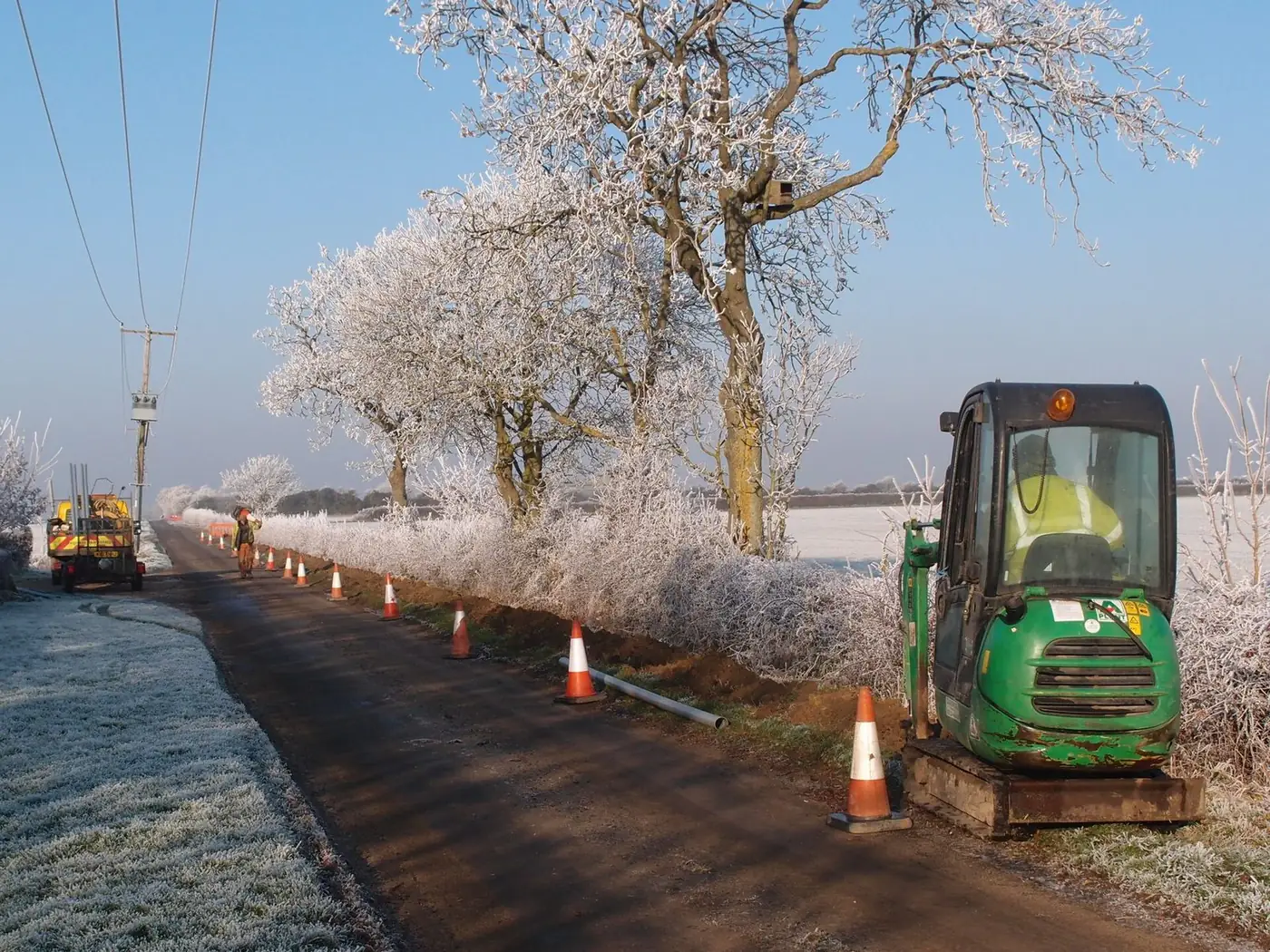 Small green mini excavator digging a trench along a roadside, with traffic cones and frosted trees