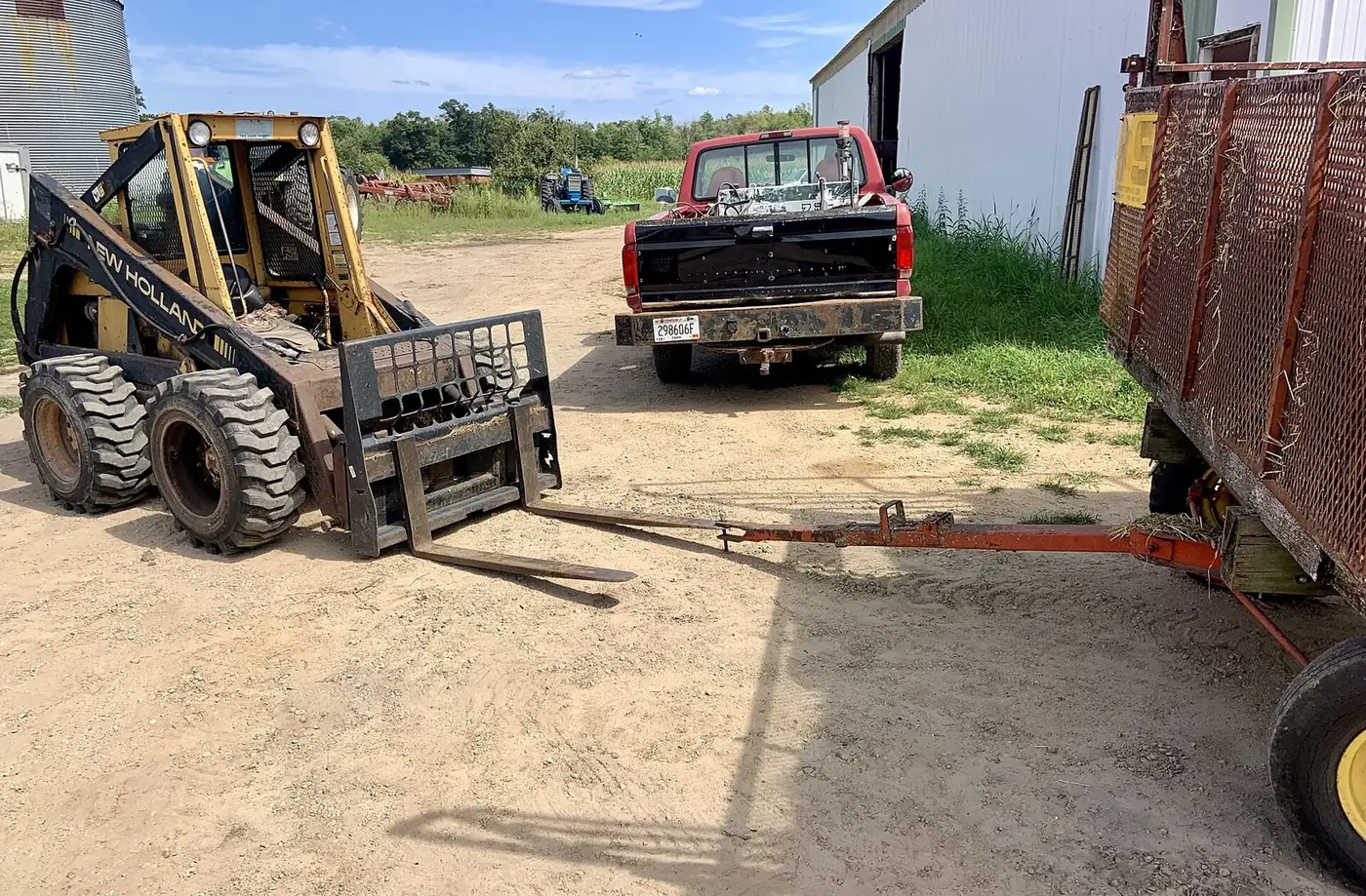 Yellow skid steer loader with forks parked on a dirt yard beside a pickup and trailer