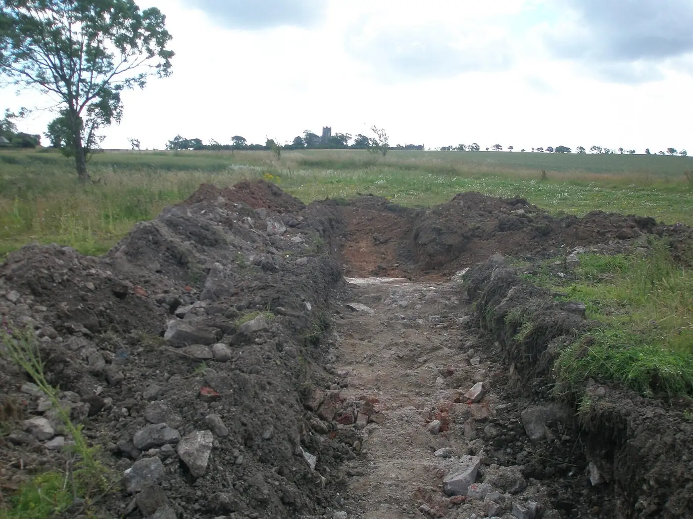A long narrow trench cut through grass and stony soil, with the spoil heaped along both sides